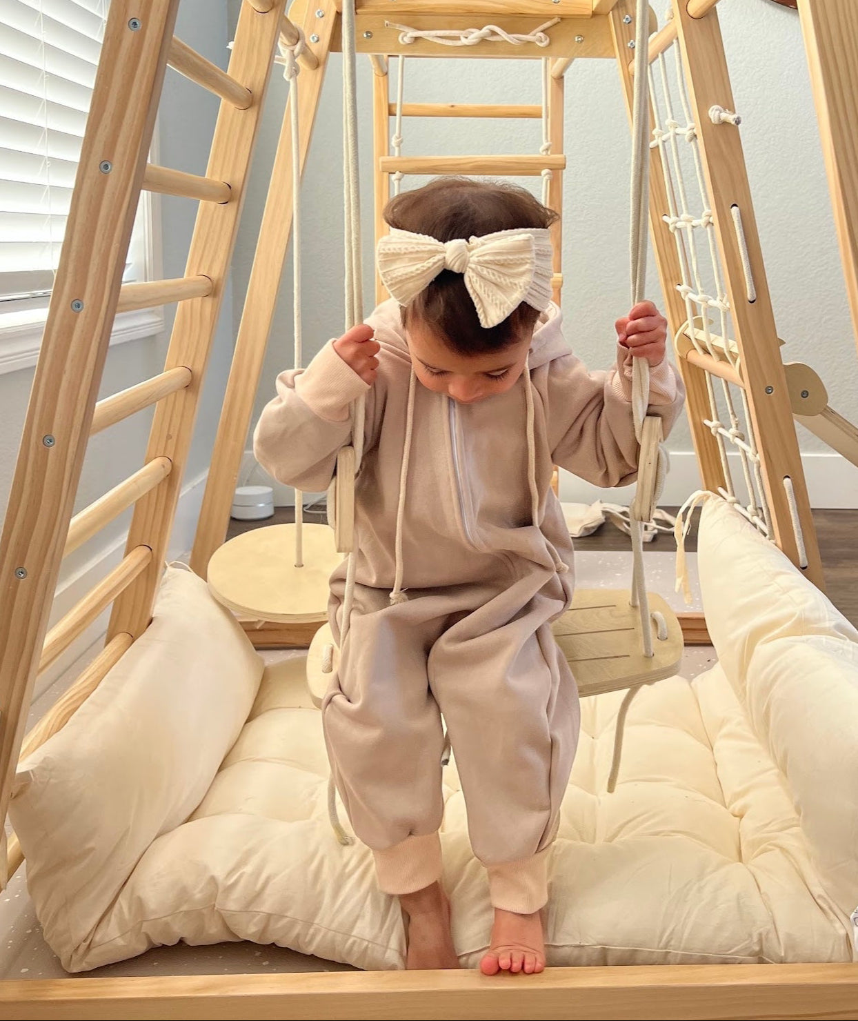 Child in a beige outfit standing on a wooden climbing frame with a swing.