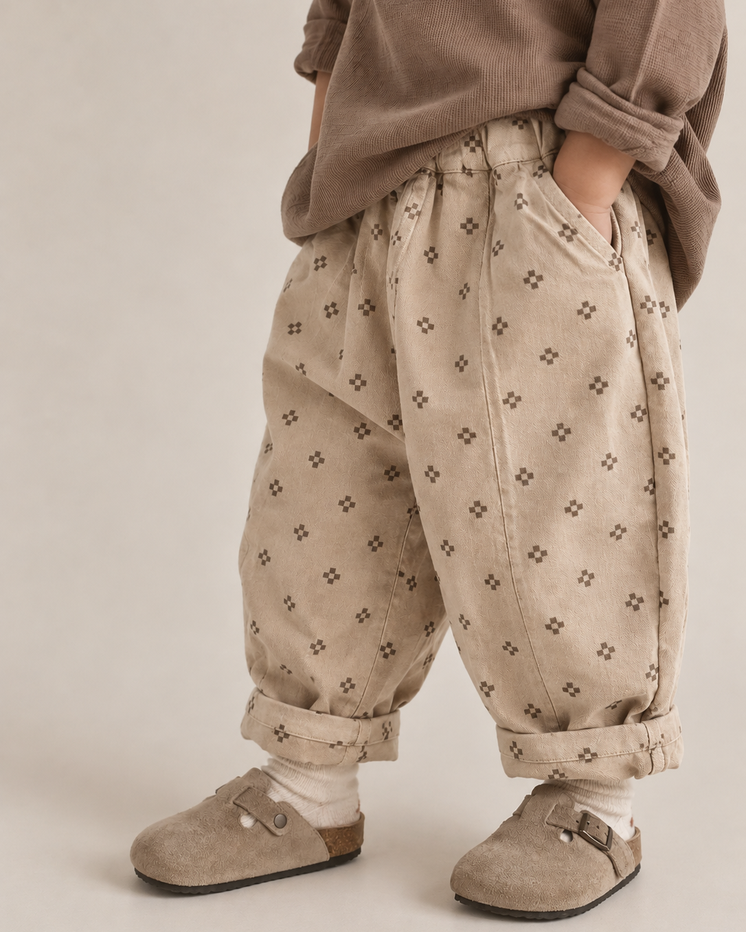 A child wearing loose beige corduroy pants with a small dark geometric pattern, styled with a brown top and brown slip-on shoes against a neutral studio background.
